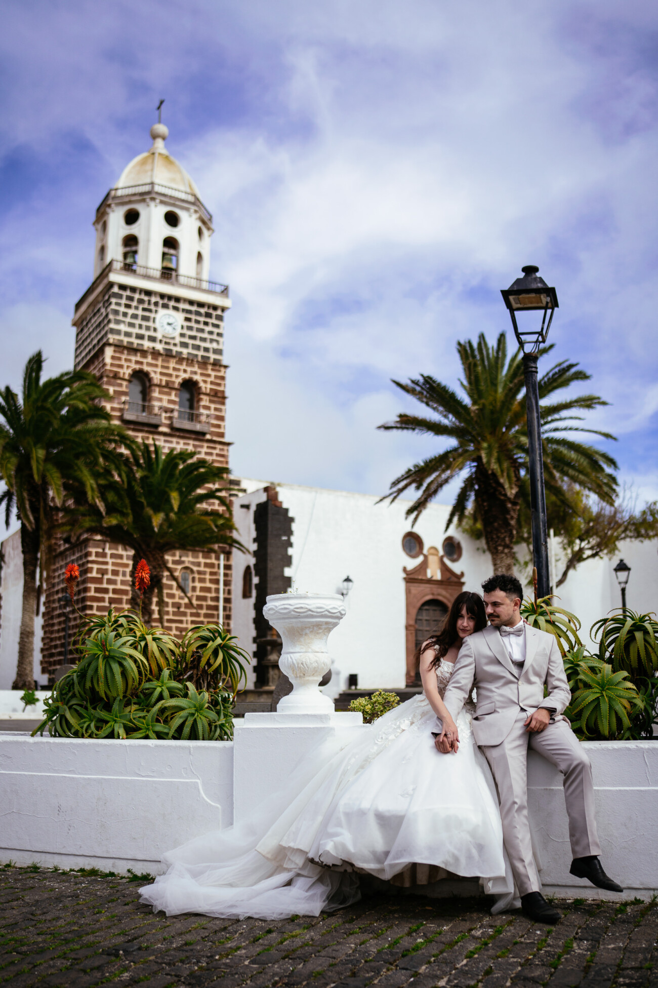 A bride and groom in formal attire pose near a white wall with greenery, showcasing destination wedding photography in front of a historic stone church tower with palm trees beneath a partly cloudy sky.
