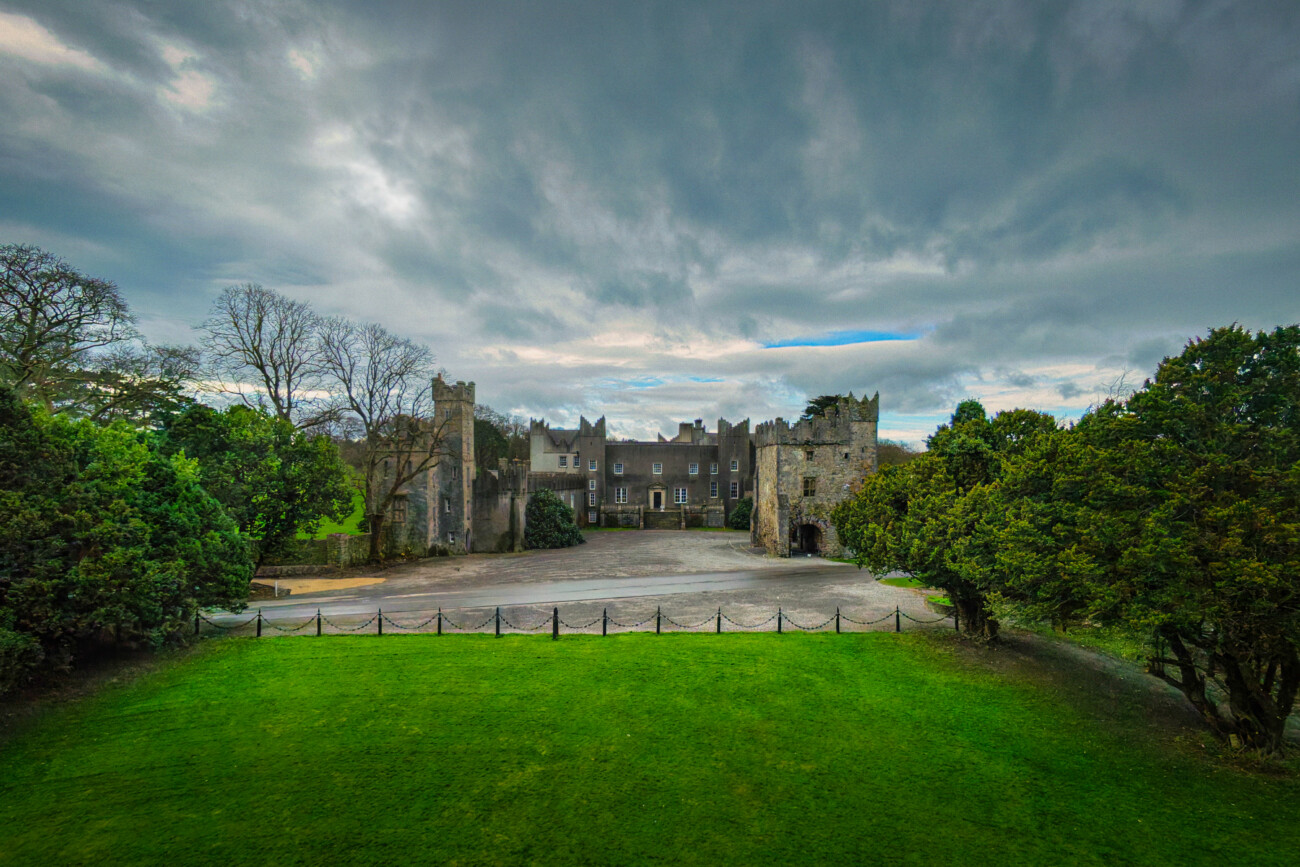 A large stone castle with towers and arched entryways sits behind a green lawn, surrounded by trees beneath a cloudy sky.