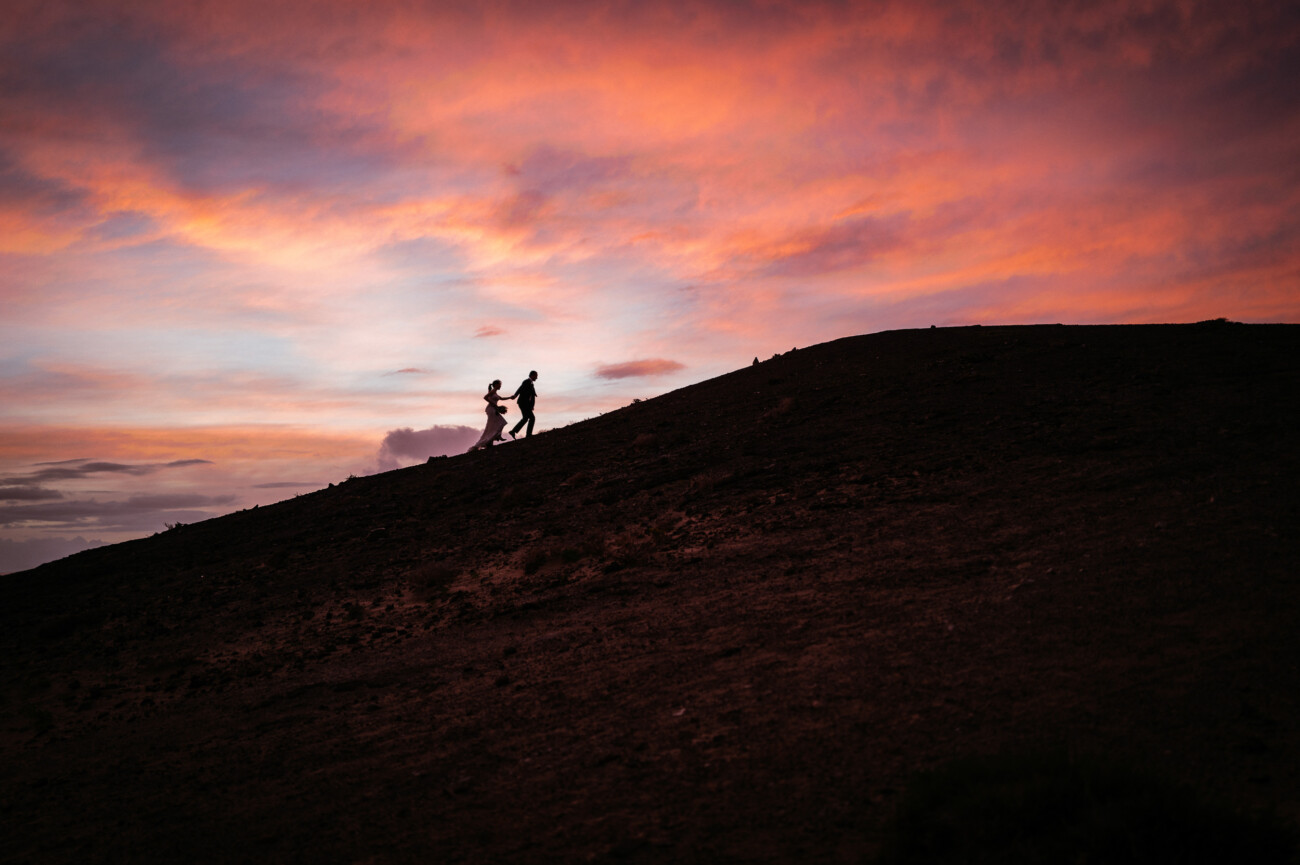 Two people walk up a hill at sunset, silhouetted against a colourful sky with shades of orange, pink, and purple—a perfect moment for destination wedding photography.