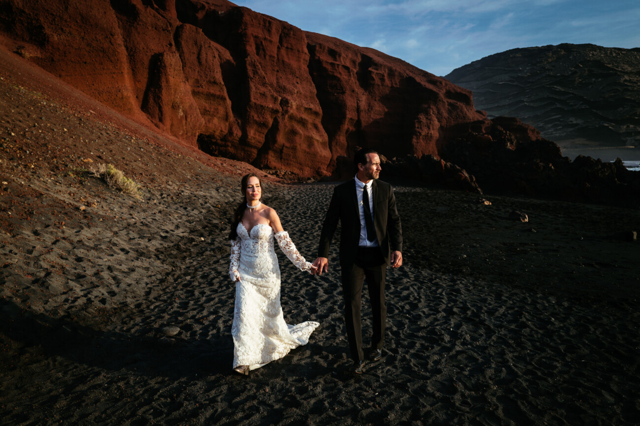 A couple in wedding attire walks hand in hand on a black sand beach with large red rock formations in the background, capturing a stunning moment of destination wedding photography.