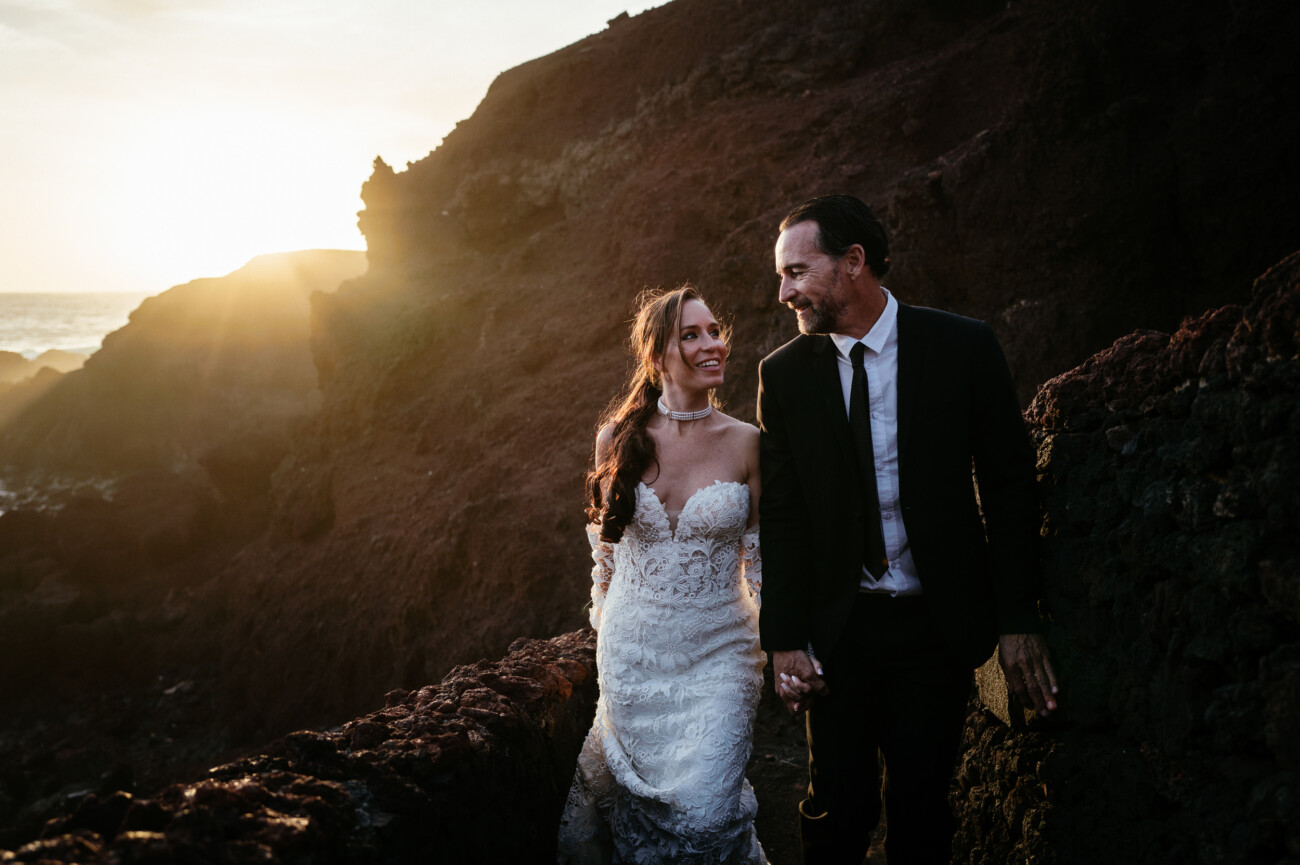 A bride in a lace gown and a groom in a suit walk hand in hand along a rocky path at sunset, capturing the romance of destination wedding photography.