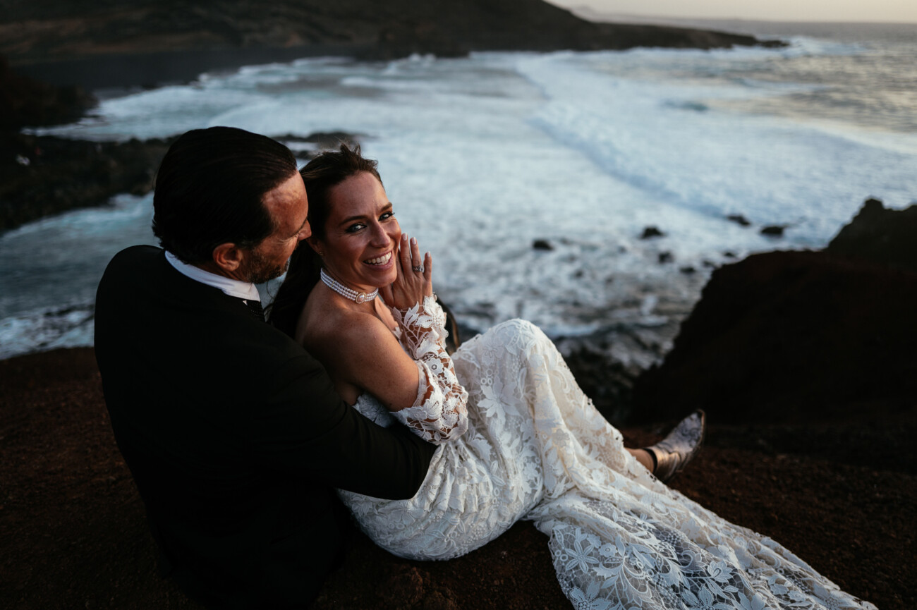 A couple in wedding attire sit together on a rocky cliff overlooking the sea, smiling and laughing as waves crash below—a perfect moment captured through destination wedding photography.