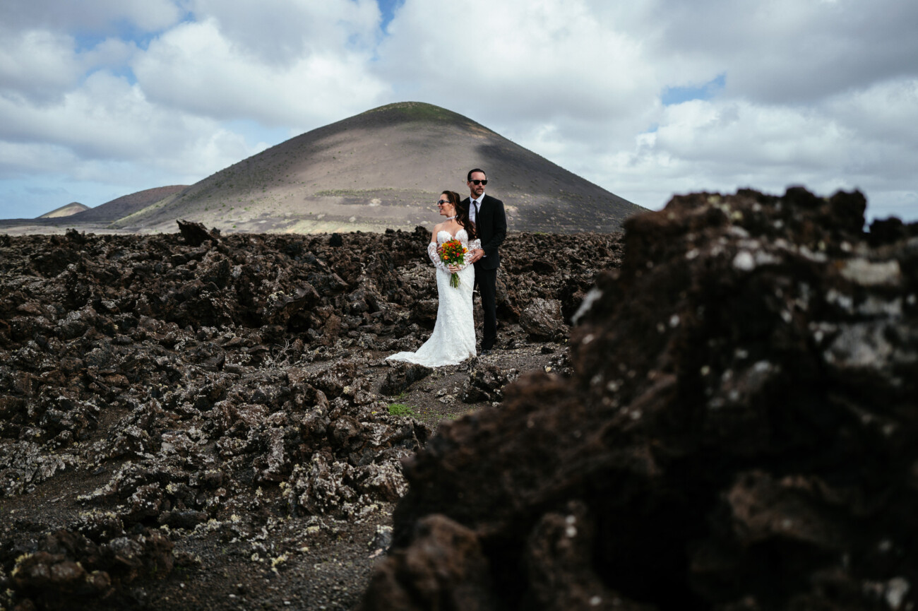 A bride and groom stand together in a rocky volcanic landscape with a mountain and cloudy sky in the background, capturing the magic of destination wedding photography.