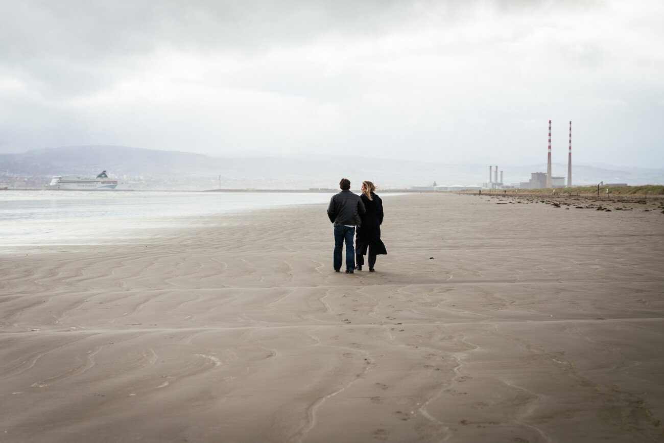 Two people walk together on a wide, empty beach with industrial buildings and smokestacks visible in the distance under a cloudy sky.