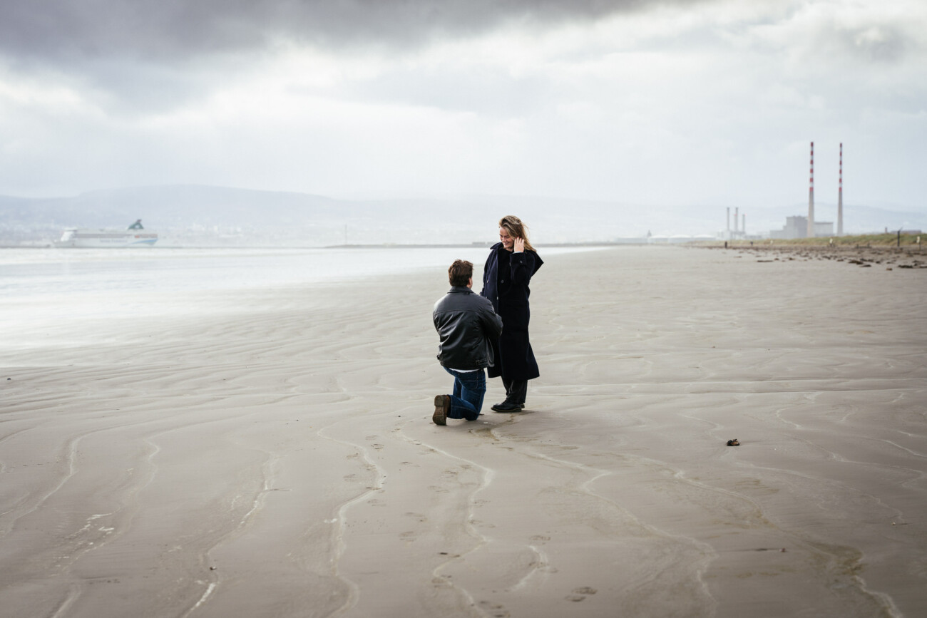 A guy kneels in front of his girlfriend on a deserted beach under cloudy skies, with industrial buildings and a ship visible in the background during his secret proposal shoot at Bull Island, Dublin