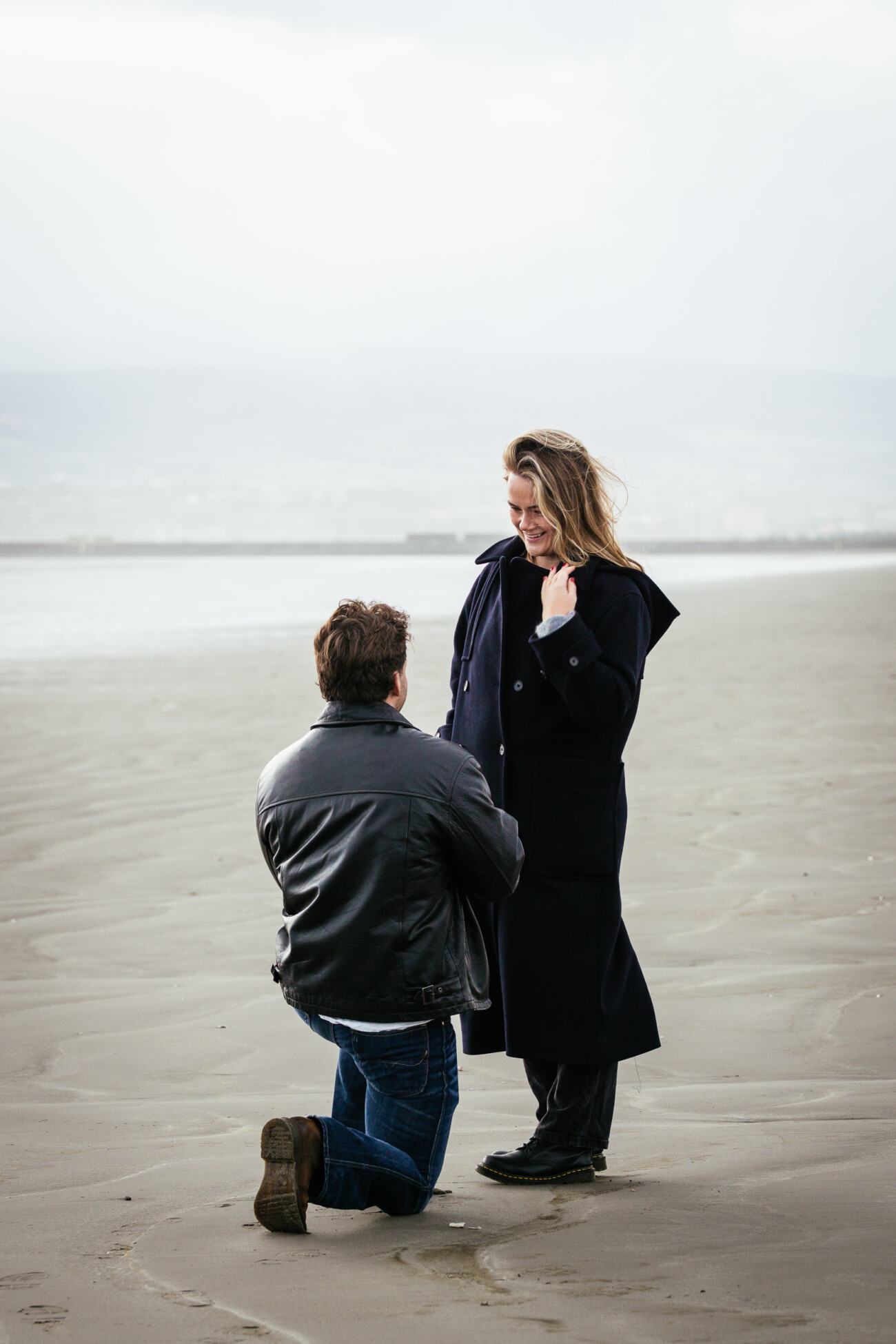 Man kneels on a sandy beach, proposing to a woman standing in front of him who appears to be smiling.