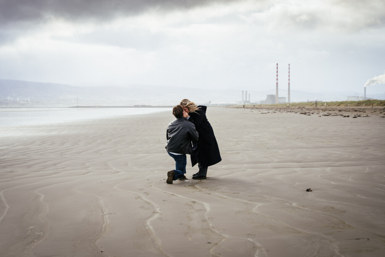 A person kneels and holds hands with another person on a deserted beach, with industrial chimneys and smoke visible in the background under a cloudy sky.