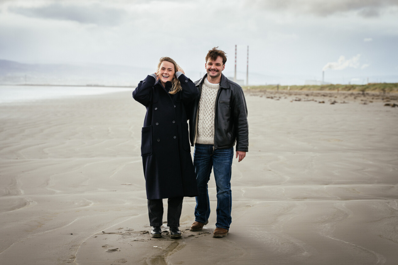 Two people standing side by side on a deserted beach, both smiling at the camera. The sky is overcast and industrial chimneys are visible in the background.