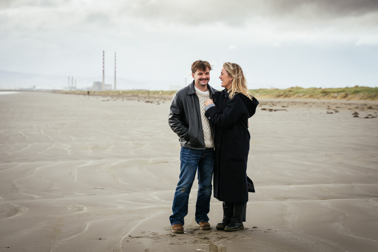 A man and woman stand together on a sandy beach under a cloudy sky, with distant industrial chimneys visible in the background.