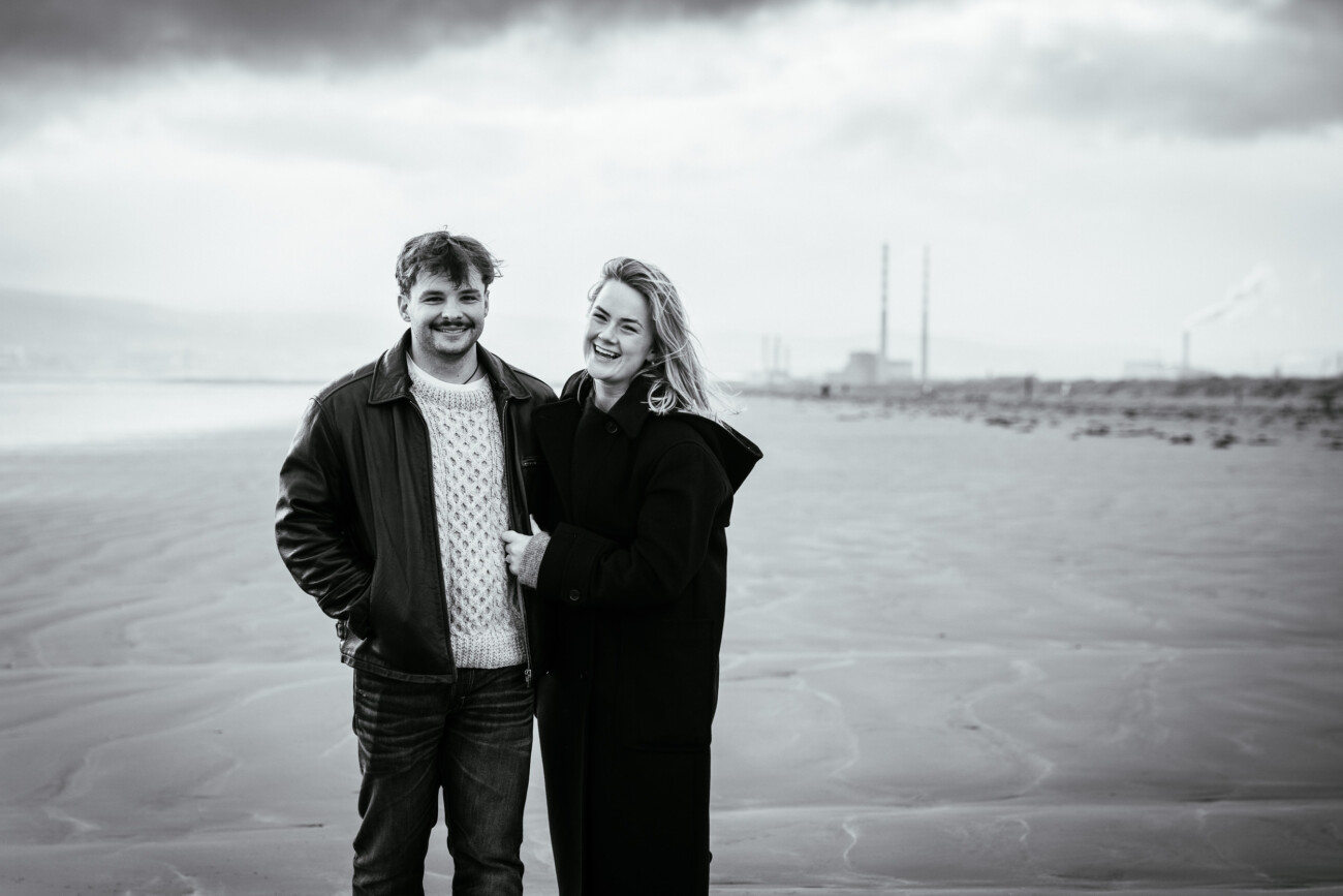 A man and woman stand together on a sandy beach, smiling at the camera. Industrial chimneys and smoke are visible in the distance under a cloudy sky.