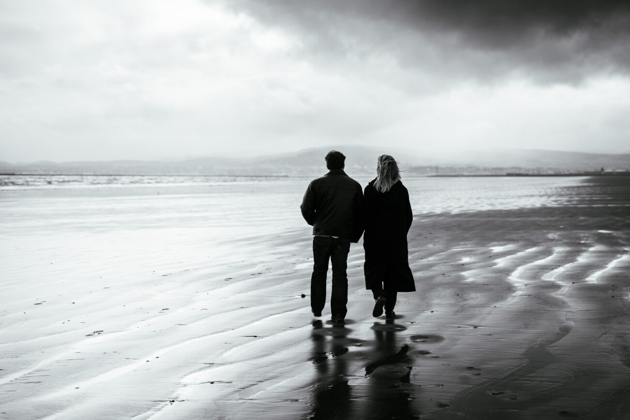 Two people walk side by side on a wet, deserted beach under a cloudy sky, with their backs to the camera.
