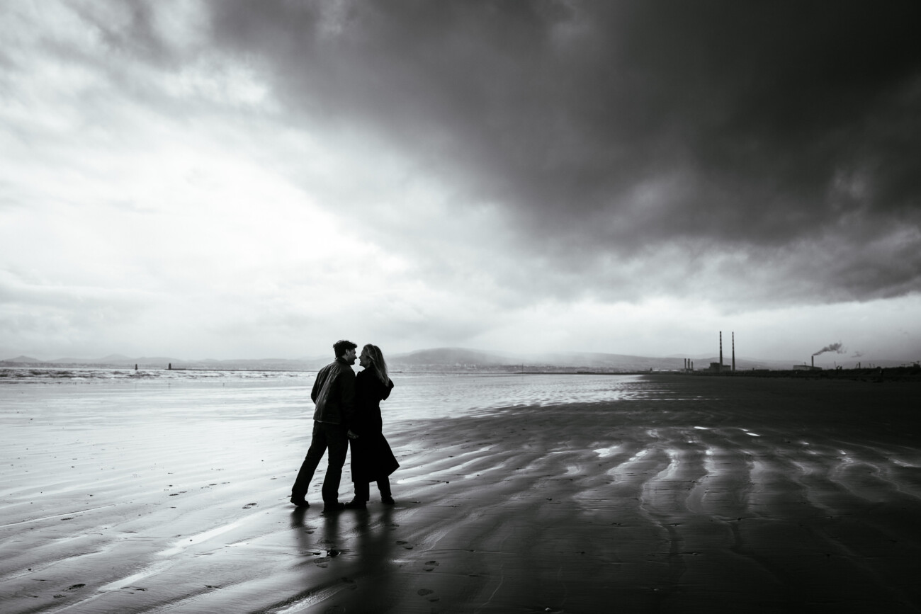 A couple stands close together on a deserted beach under a cloudy sky, with industrial chimneys visible in the distant background.