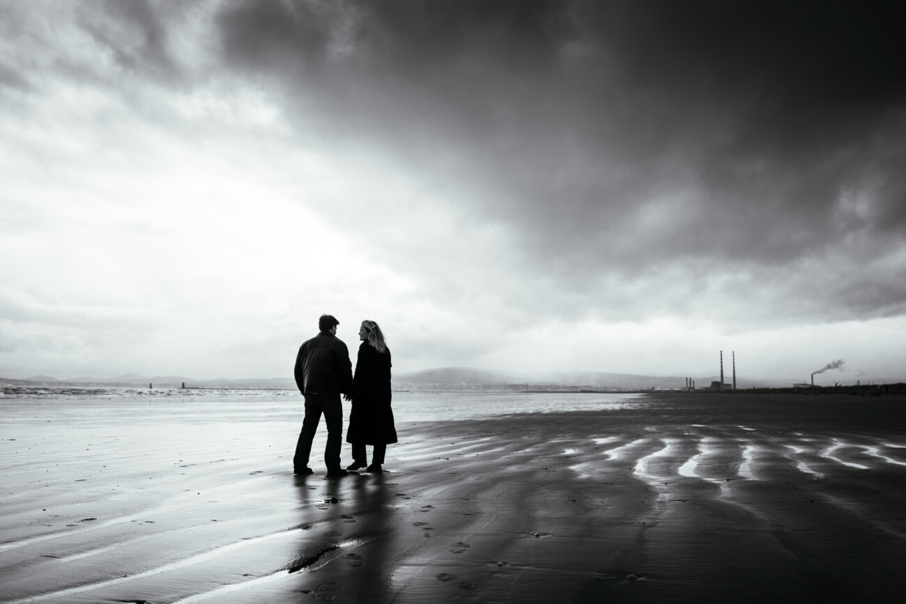 Two people stand on a wet, empty beach under a cloudy sky, facing each other, with industrial chimneys visible in the distant background.
