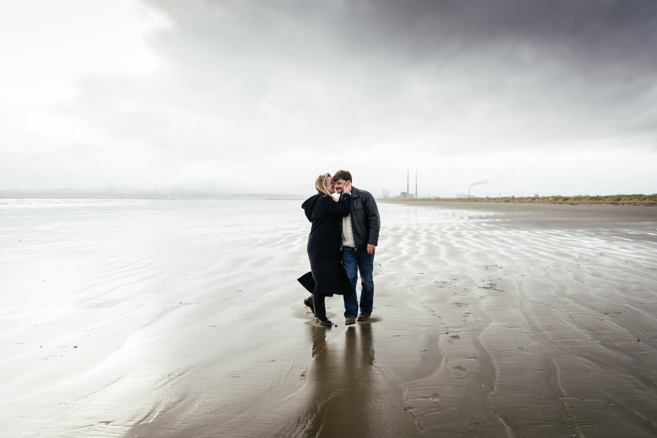 Two people walk closely together on a wet, sandy beach under a cloudy sky, with industrial chimneys visible in the background.