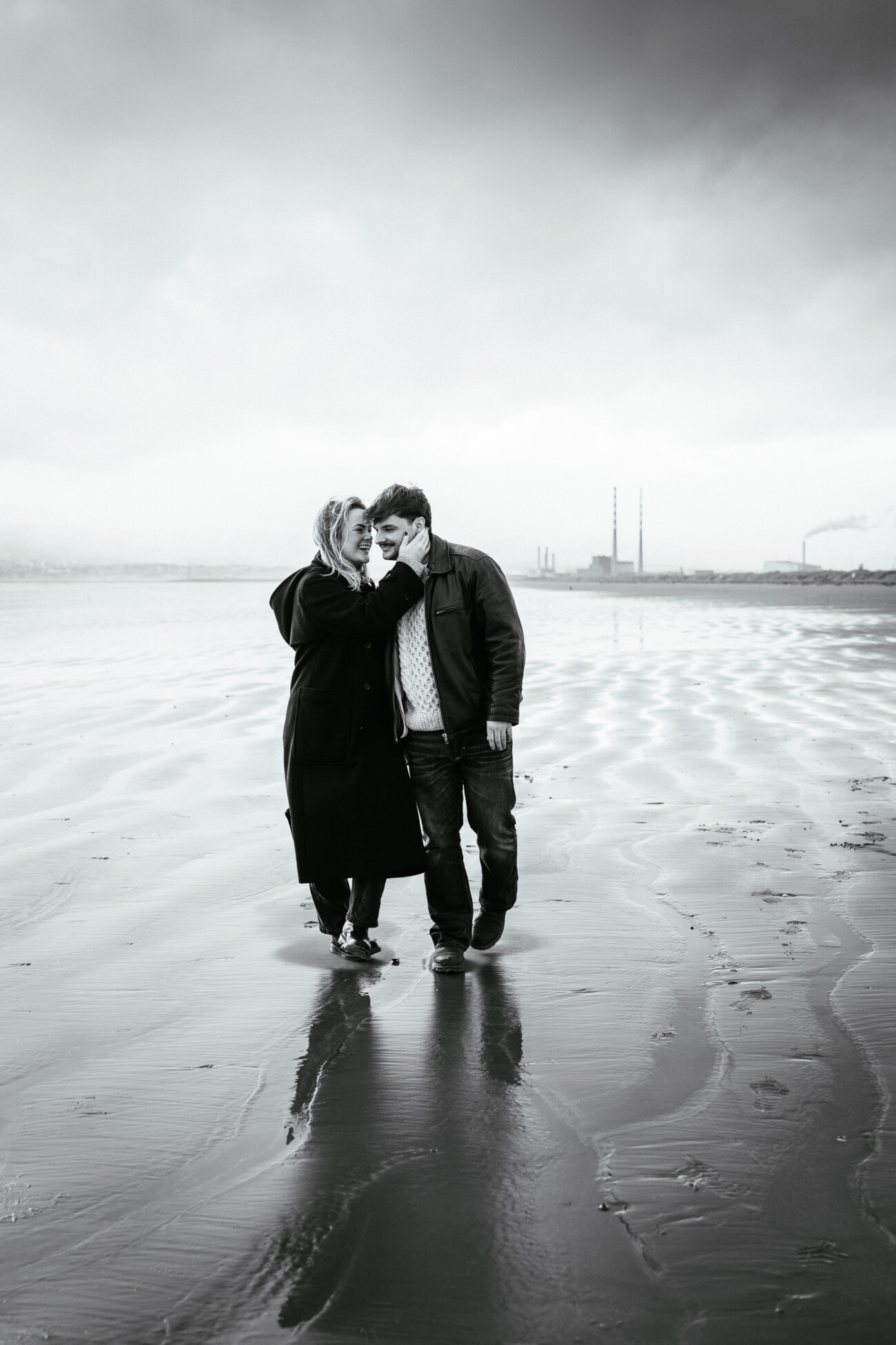 A couple walk on a wet, sandy beach; the woman touches the man's face. Industrial chimneys and a distant city skyline are visible in the background under a cloudy sky.