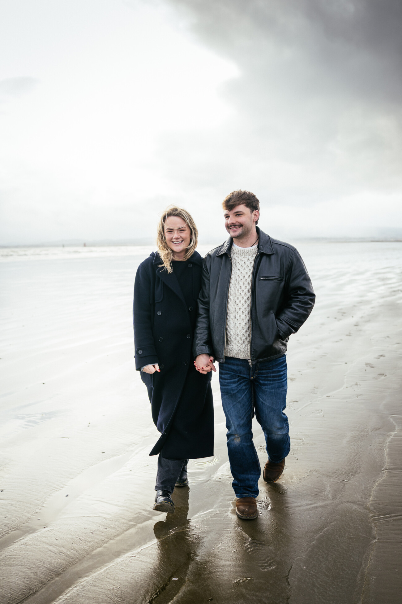A woman and a man walk hand in hand along a sandy beach on a cloudy day, both wearing jackets and smiling.