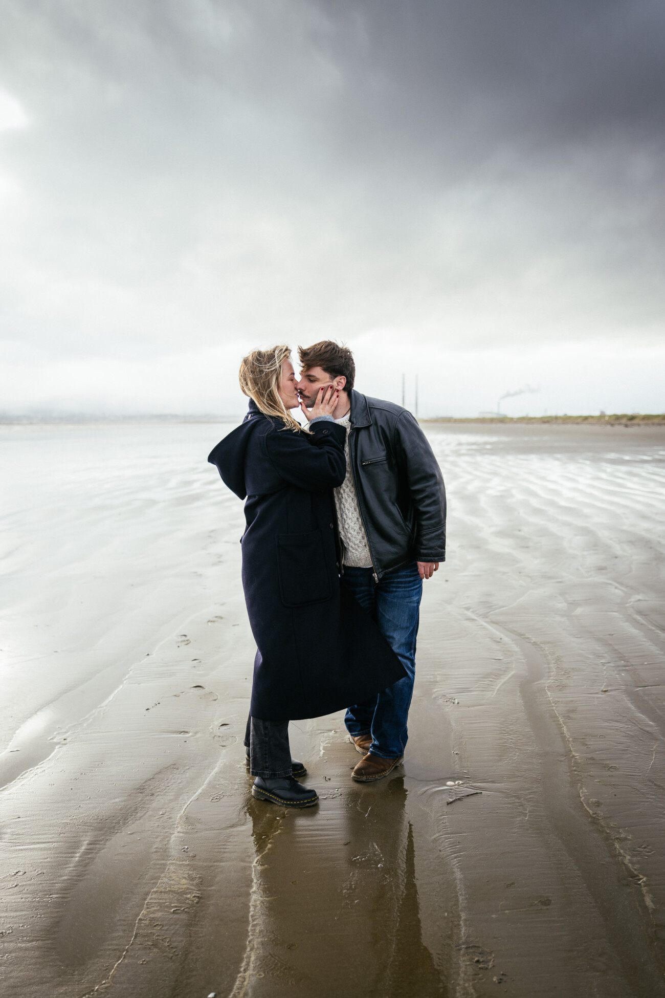 A couple stands close together on a wet, overcast beach, with the woman touching the man's face and both dressed in coats and boots.