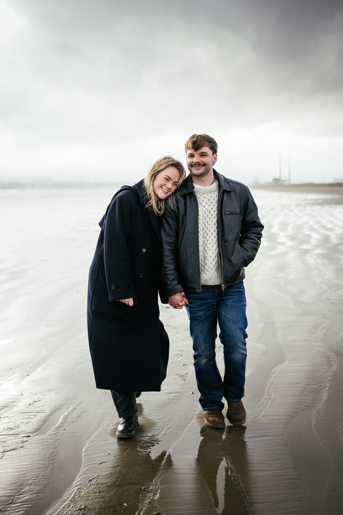 Two people stand on a wet, sandy beach holding hands, dressed in coats and jeans, with a cloudy sky and distant industrial buildings in the background.