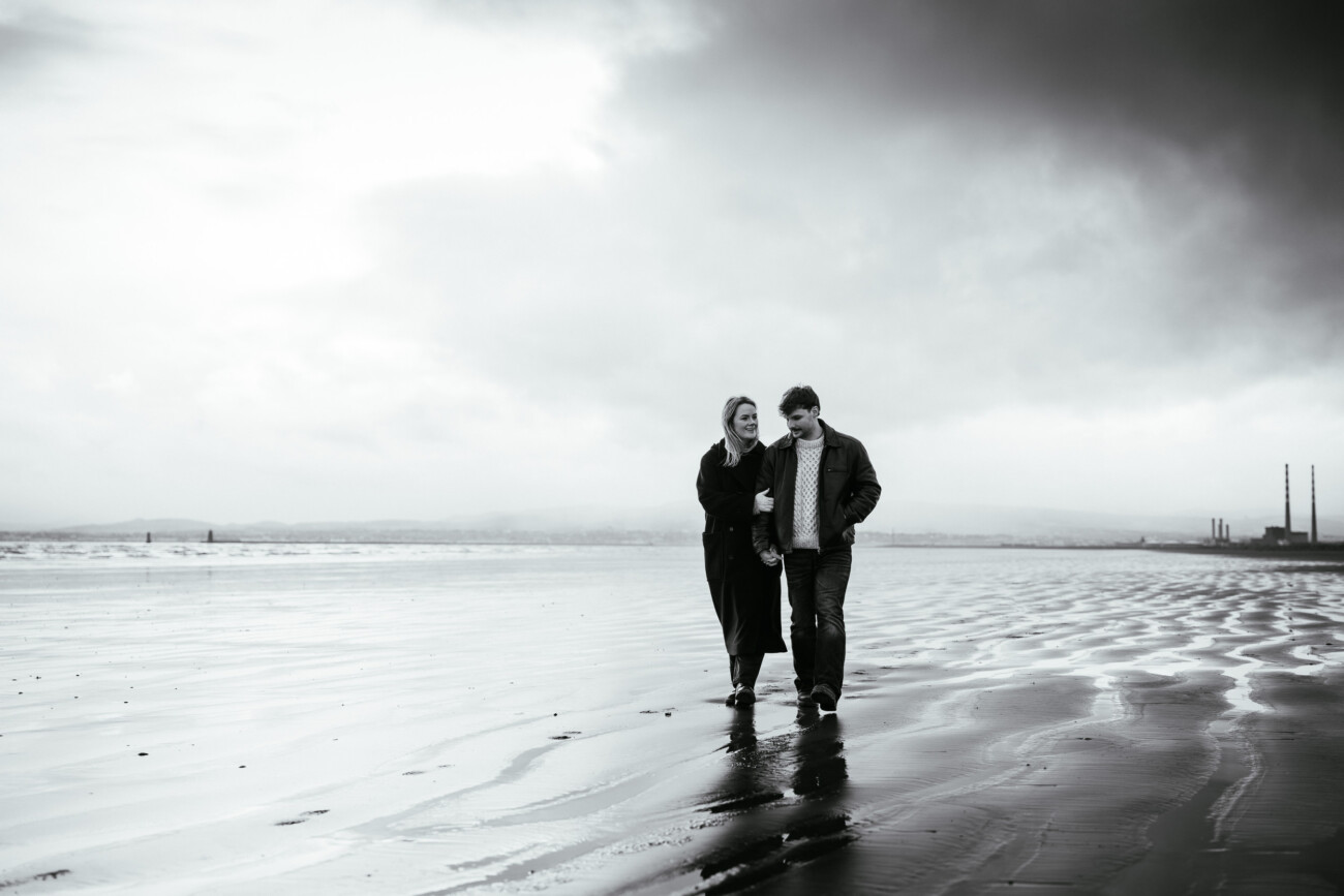Two people walk together on a wet, sandy beach under a cloudy sky, with industrial chimneys visible in the distant background.