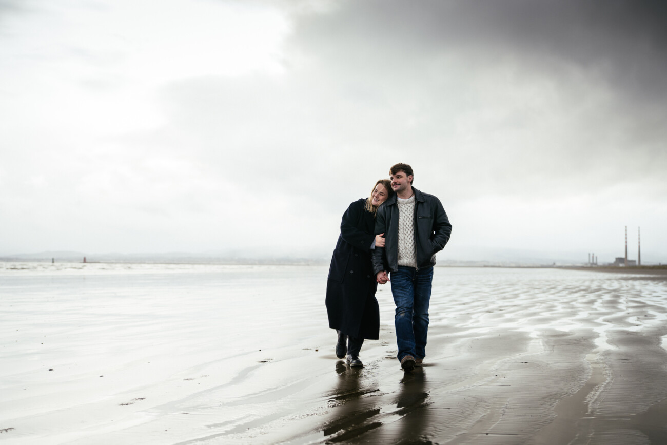 A couple walks along a wet, empty beach under a cloudy sky, holding hands and leaning into each other.