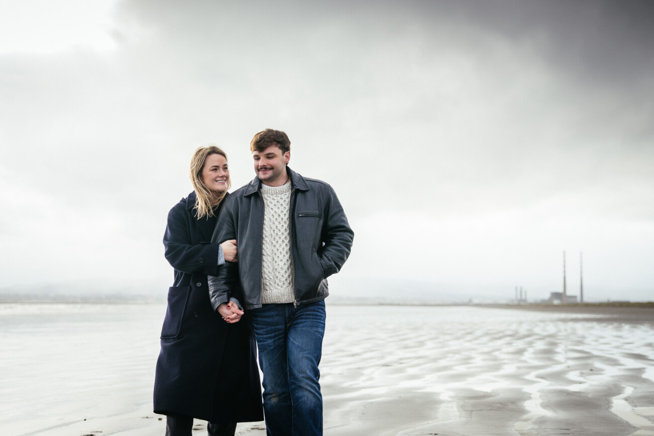 A man and woman walk together on a cloudy, deserted beach, holding hands and wearing jackets. Industrial chimneys are visible in the background.