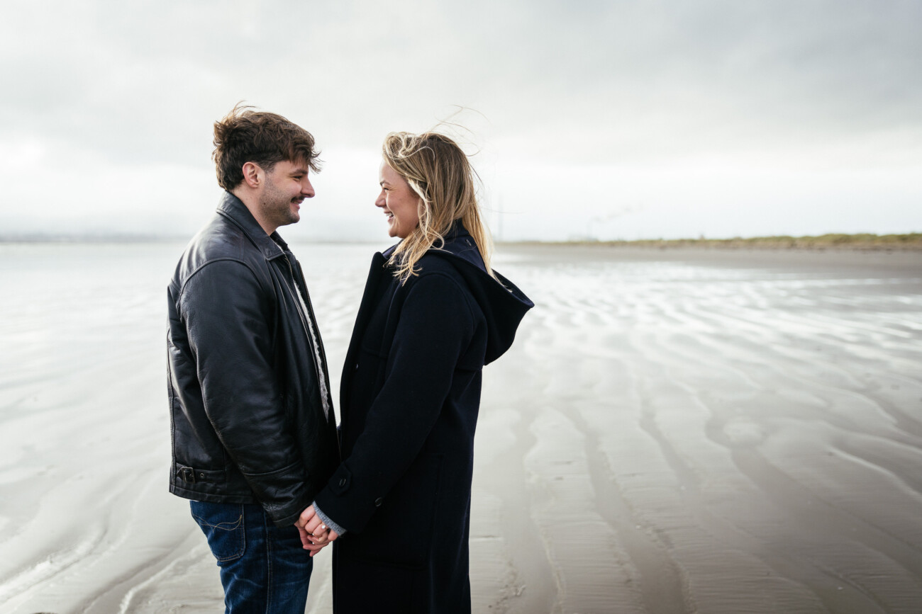 A man and woman stand facing each other, holding hands and smiling, on a cloudy beach with wet sand and distant industrial structures in the background.