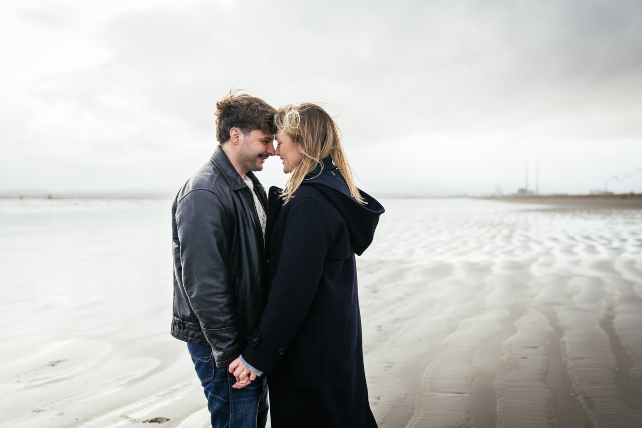 A man and woman stand close together, holding hands and touching foreheads, on a cloudy, deserted beach with rippled sand.