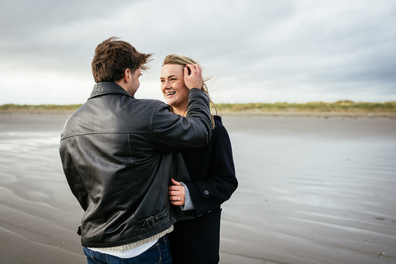 A man and woman stand on a beach; the man faces away, touching the woman's hair, whilst the woman smiles and looks at him. The beach is empty with overcast skies.