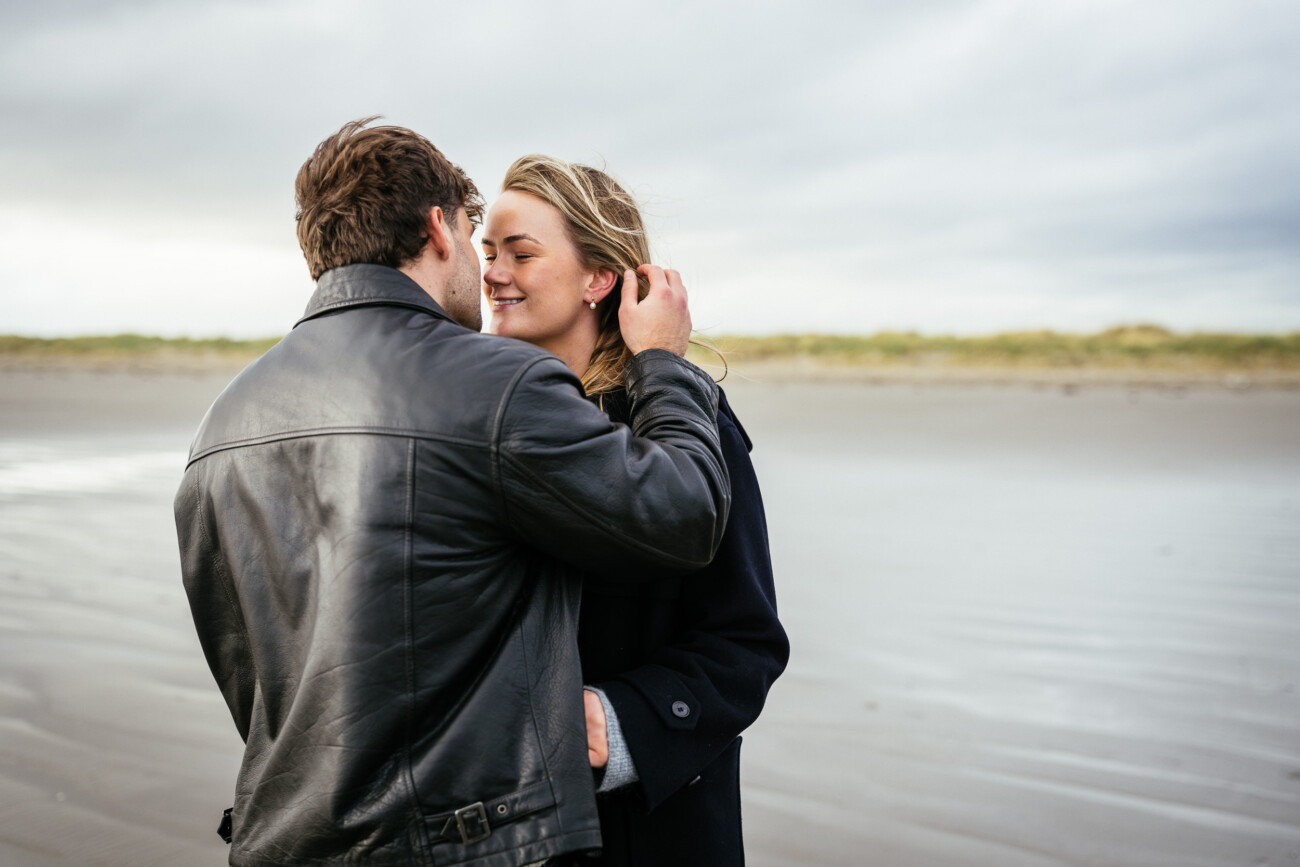 A man and woman stand close together on a sandy beach, facing each other affectionately under an overcast sky.