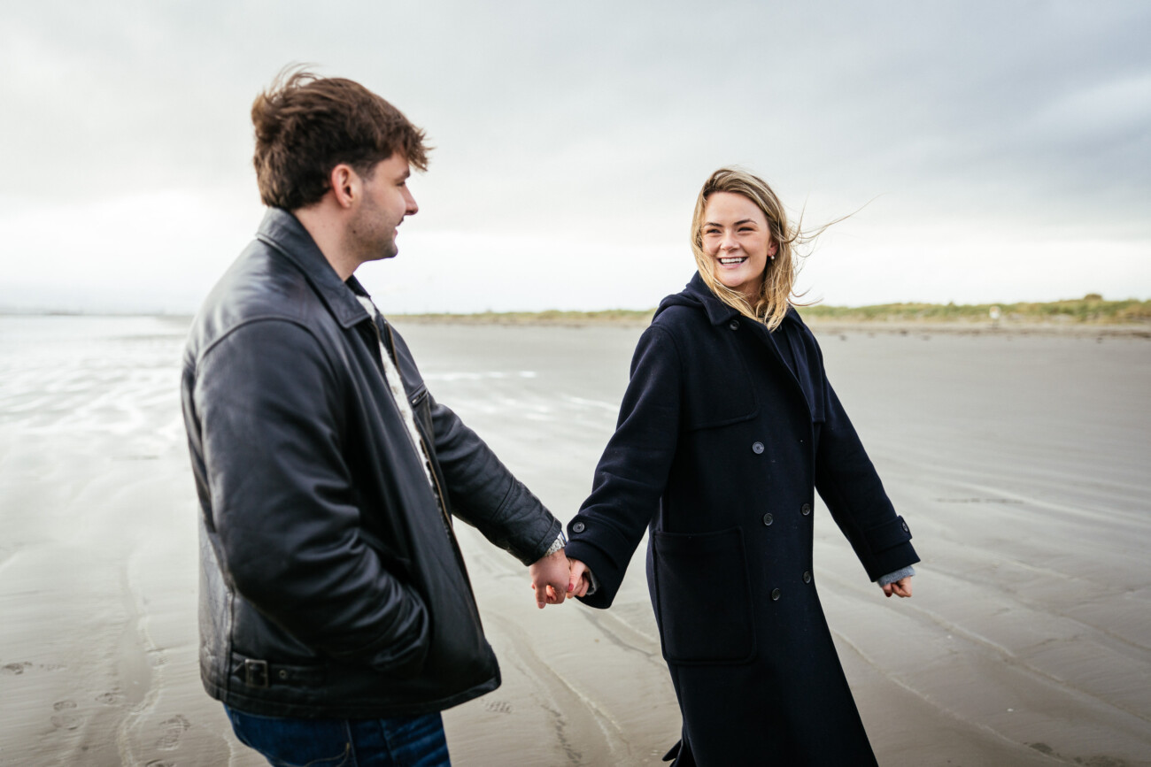 Two people holding hands and walking on a sandy beach, both wearing coats. The woman is smiling whilst looking at the man. The sky is overcast.