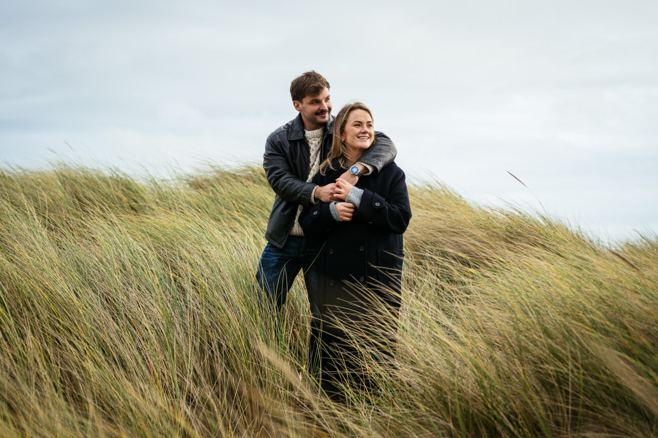 A man stands behind a woman in tall grass, his arms round her shoulders. Both are wearing coats and smiling, with cloudy sky in the background.