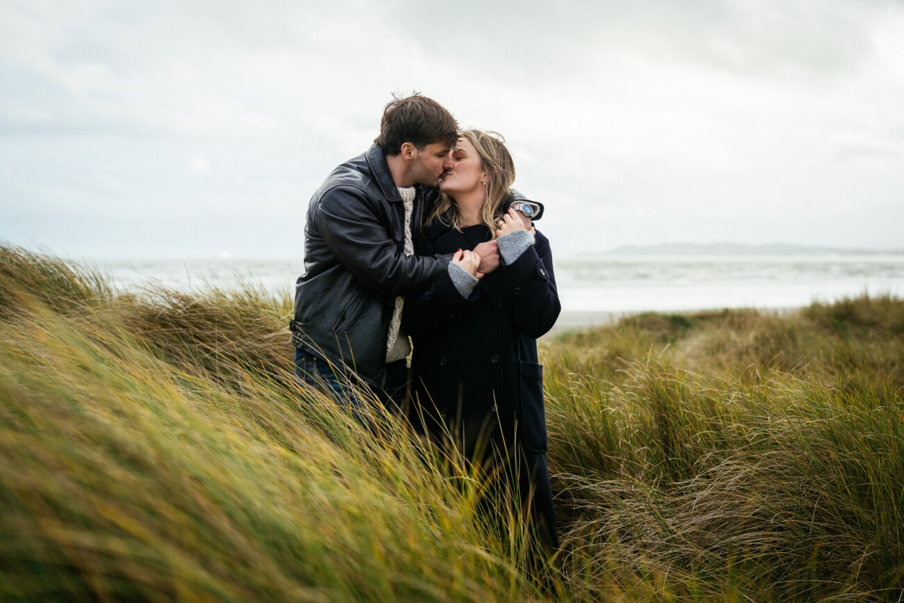 A couple stands close together in tall grass by the beach, sharing a kiss on a cloudy day with the sea in the background.