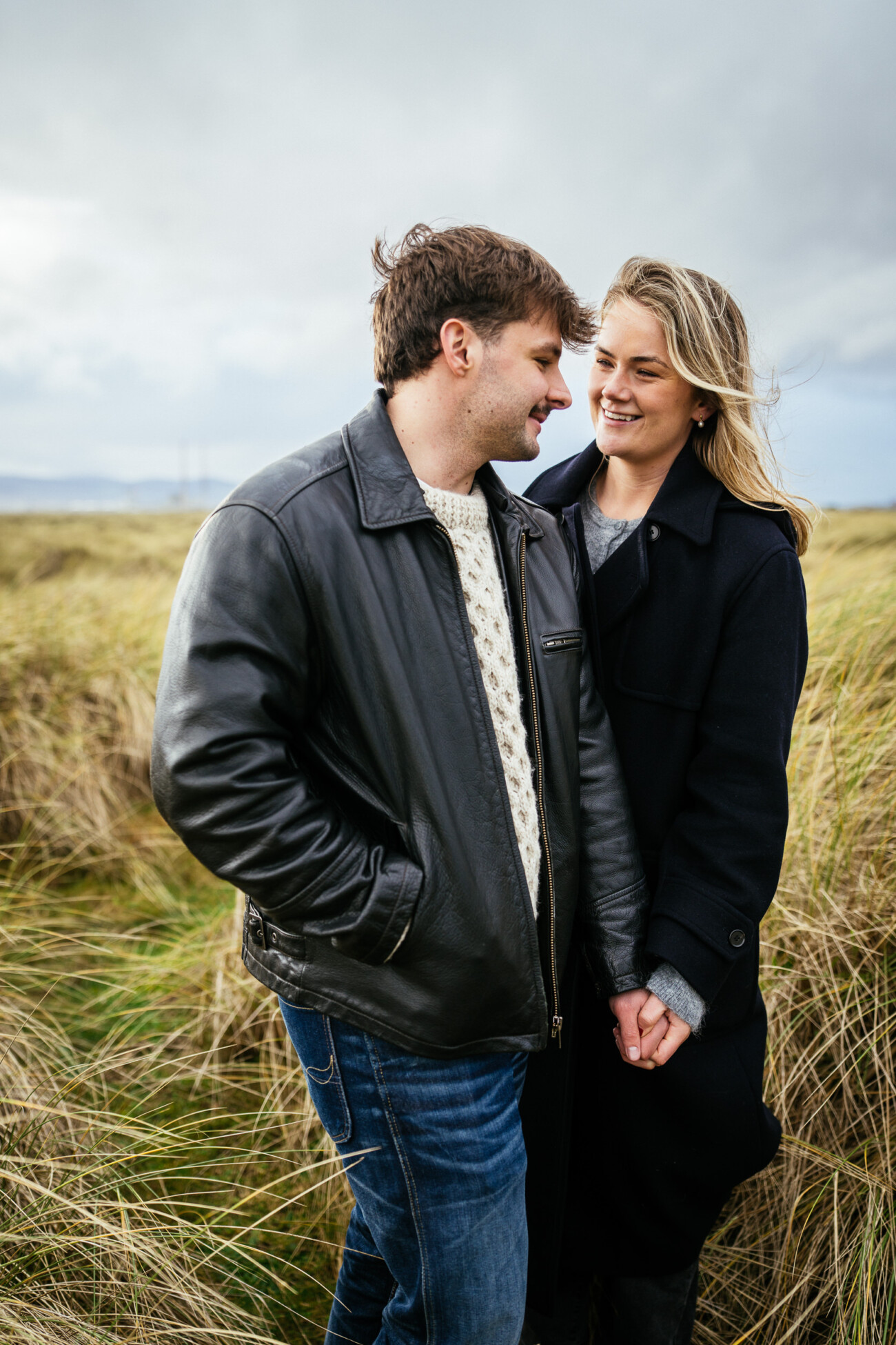 A man and a woman stand close together in a grassy field, holding hands and smiling at each other under a cloudy sky.