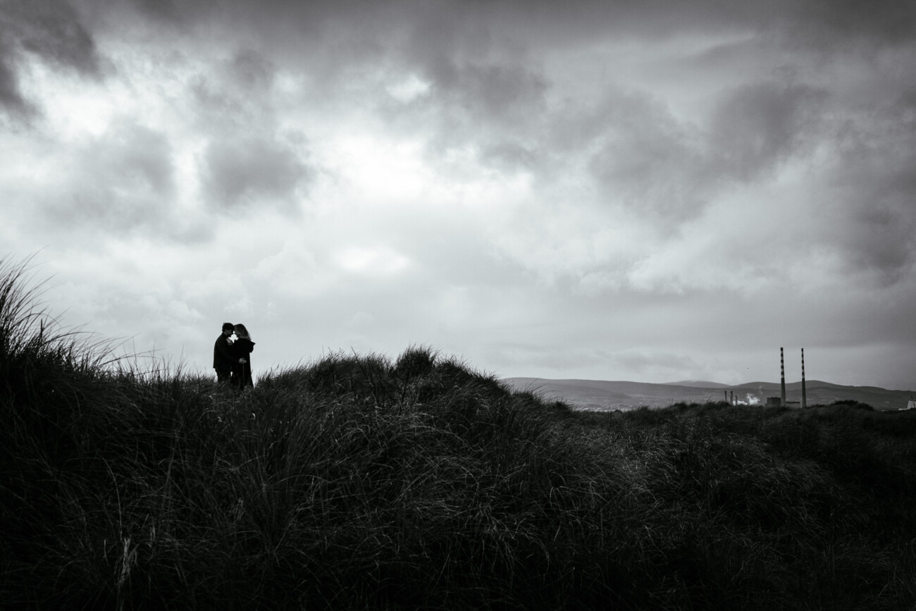 Two people stand together on a grassy hill under a cloudy sky, with distant hills and telegraph poles visible in the background.