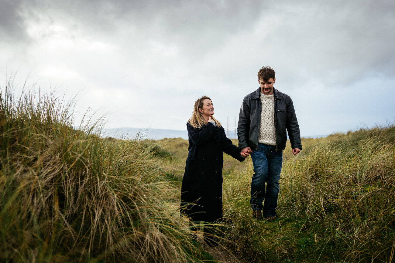 A man and a woman walk hand in hand along a grassy path under a cloudy sky, both wearing coats and looking at each other.