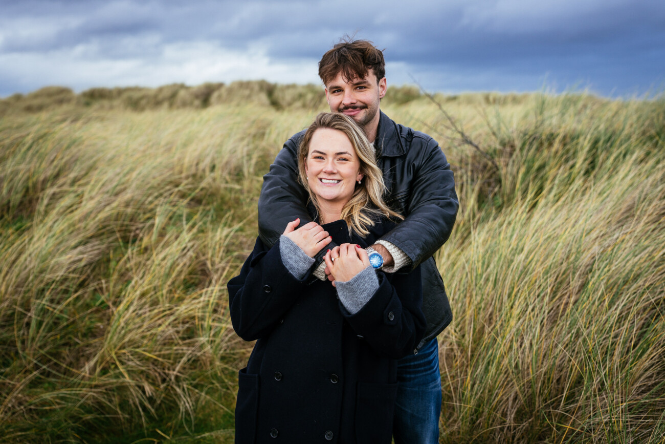 A man stands behind a woman, both smiling, as they pose together outdoors in a grassy field under a cloudy sky.