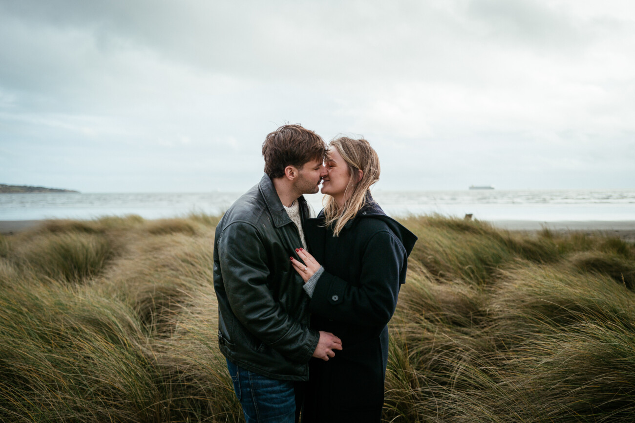 A couple stands close together, sharing a kiss in front of tall grass on a beach with cloudy skies and the sea in the background.