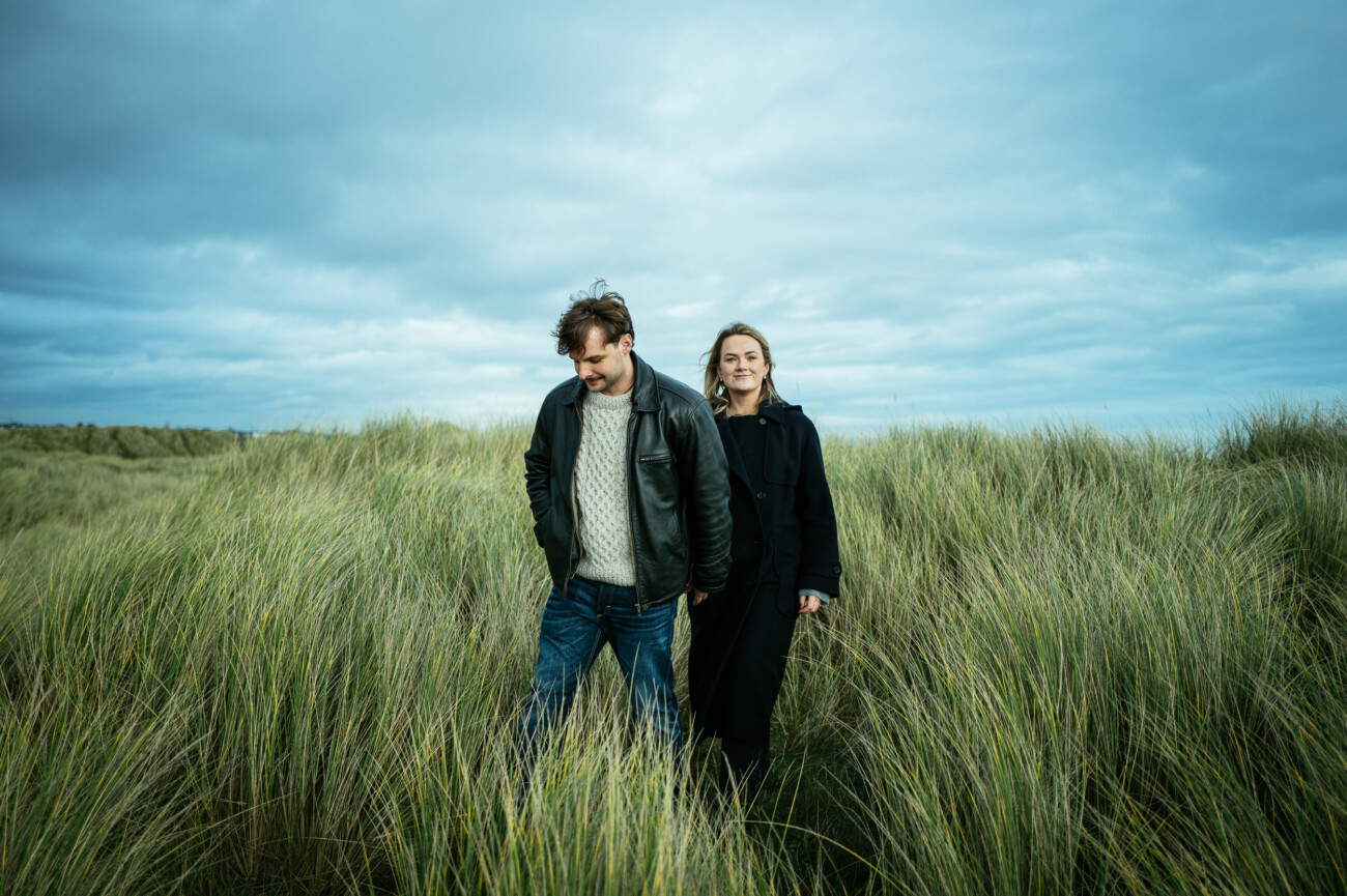 Two people stand in tall grass under a cloudy sky, one wearing a jacket and jeans, the other in a long dark coat, both looking relaxed.