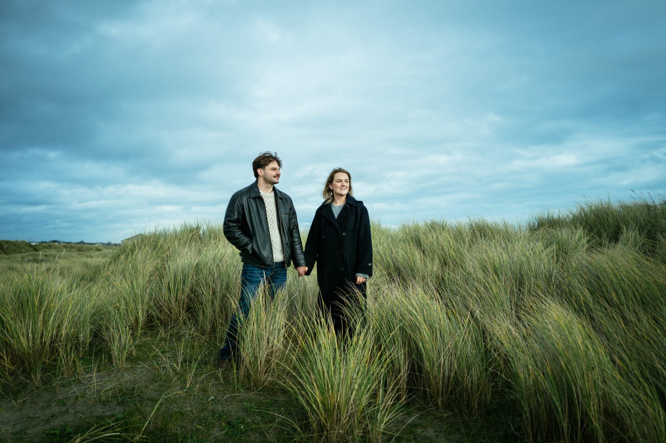 Two people stand holding hands in tall grass under a cloudy sky, both wearing dark jackets and looking off into the distance.