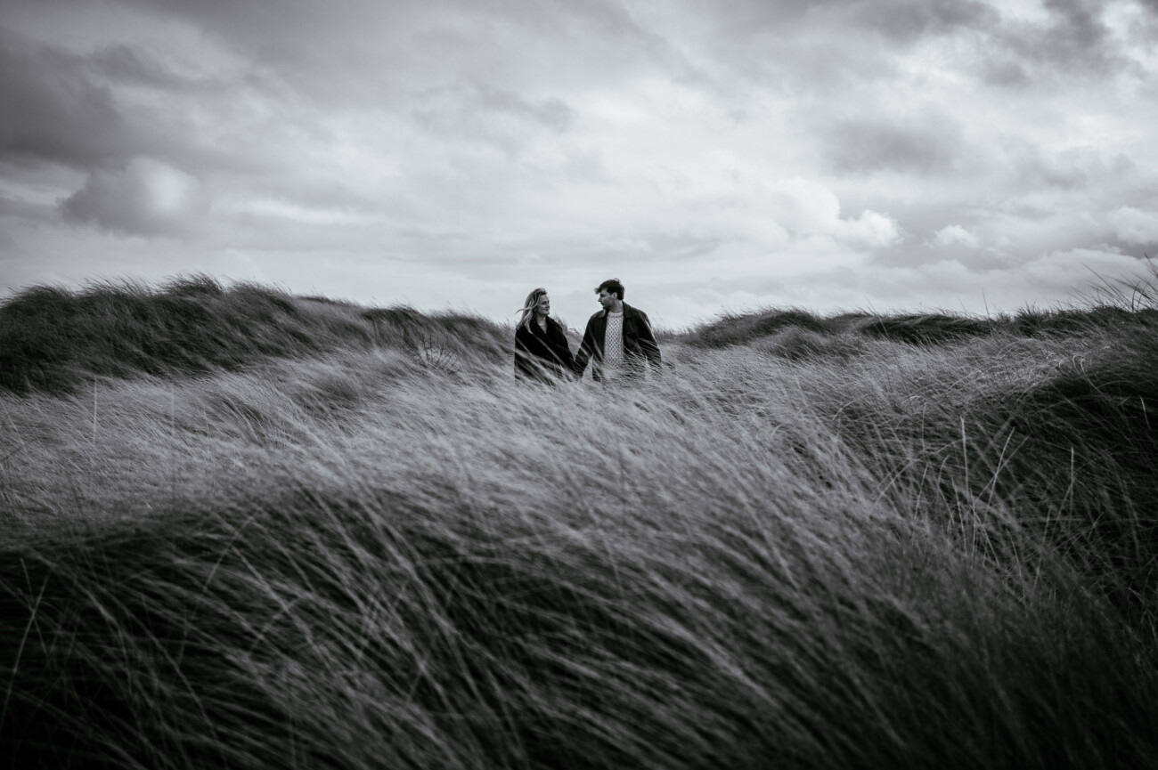Two people walk hand in hand through tall grass on a windy, overcast day, surrounded by rolling dunes under a cloudy sky.