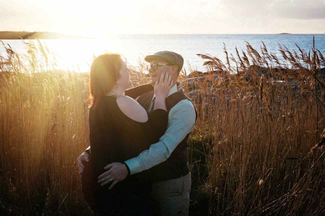 A couple stands in tall grass by a body of water at sunset, smiling and facing each other as one person touches the other's cheek—captured beautifully by a Proposal & Engagement Photographer on the wild atlantic way in Ireland.