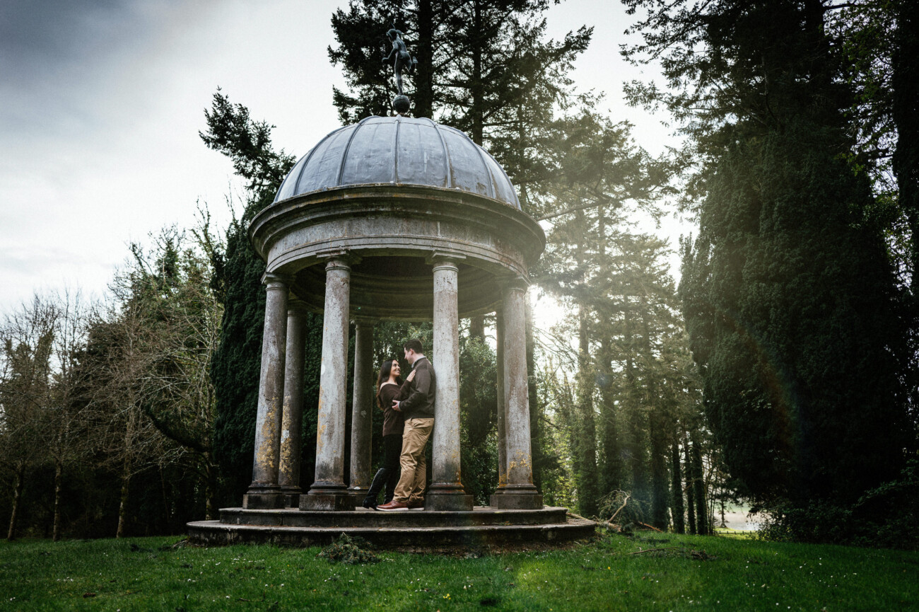 A couple stands close together inside a small stone gazebo with columns, surrounded by tall trees and greenery, during their Dromoland Castle Engagement Shoot.