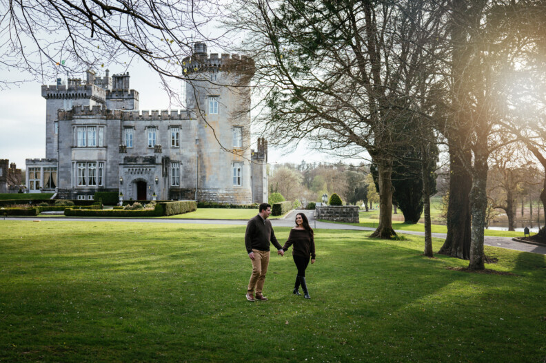 Golden Hour & Castle Magic: A Dromoland Castle Engagement Shoot 1 A couple holding hands walks on a grassy lawn in front of Dromoland Castle, surrounded by trees on a sunny day—capturing the magic of a perfect engagement photoshoot.