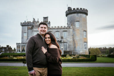 A couple stands close together and smiles in front of the grand Dromoland Castle on a cloudy day, capturing a perfect Dromoland Castle engagement photoshoot moment.