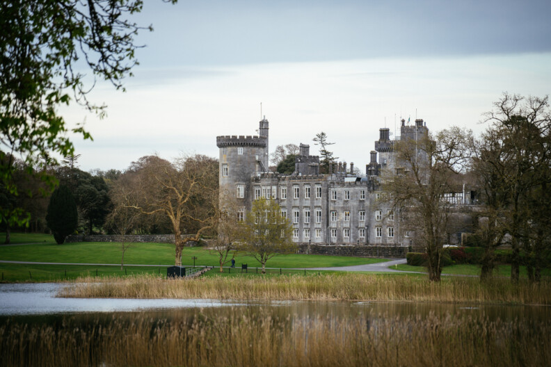 Dromoland Castle, Co Clare with turrets stands beside a lake, perfect for an engagement shoot, surrounded by lawns and trees beneath a cloudy sky.