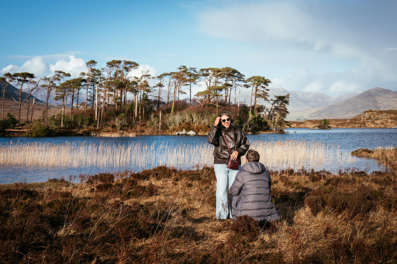A guy kneels in front of another by a serene lake, surrounded by trees and mountains—perfect for a magical proposal, beautifully captured by an engagement photographer in Connemara National Park, Ireland.