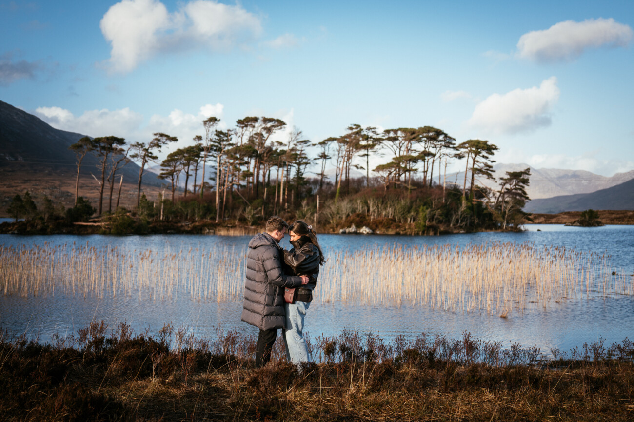 A couple stand close together by a lake with tall reeds, an island of trees, and mountains behind them under a blue sky—captured perfectly by a proposal & engagement photographer in Connemara National Park, Ireland .