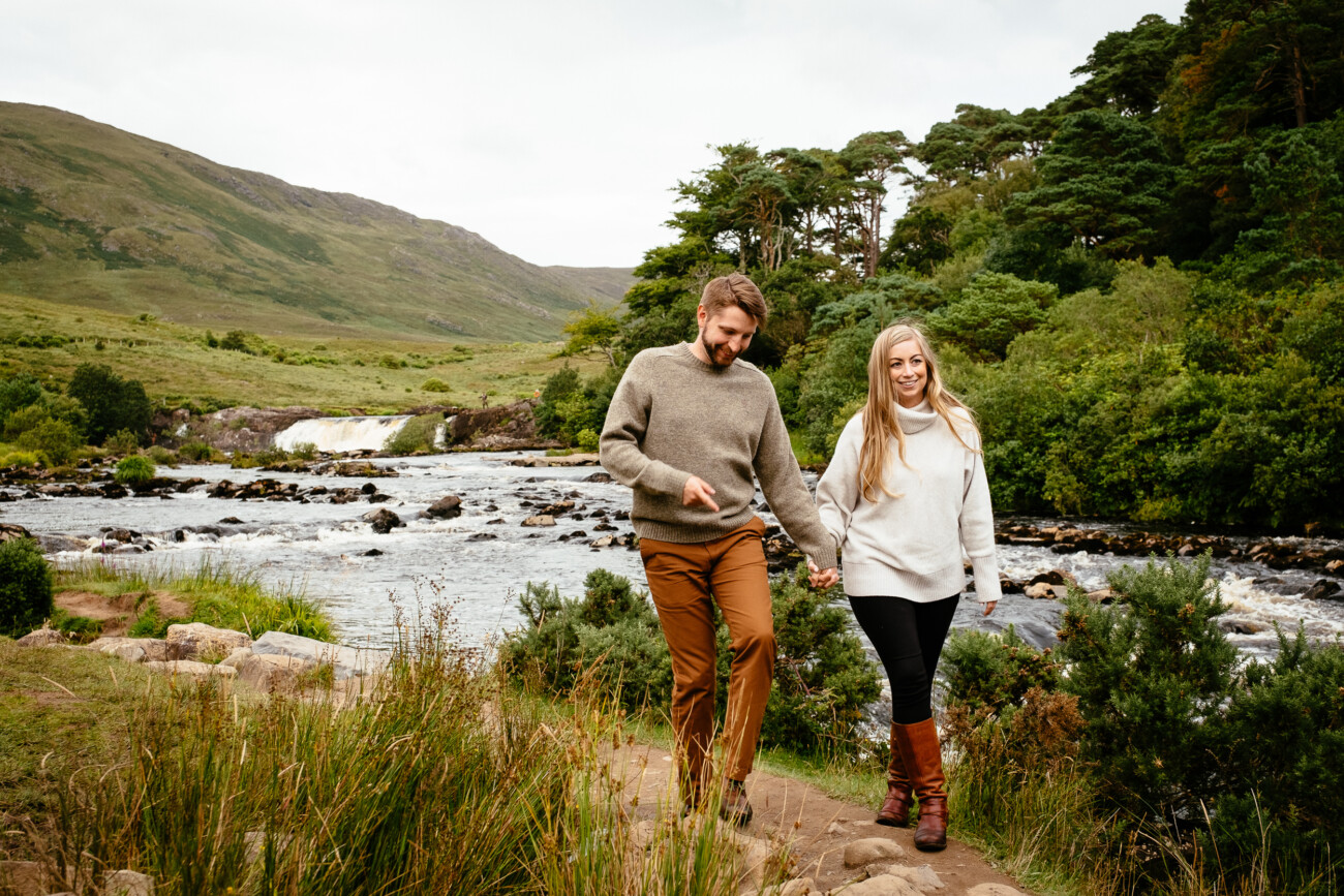 A man and woman walk hand in hand along a riverside trail with trees, hills, and a small waterfall in the background—captured by a Proposal & Engagement Photographer Ireland.