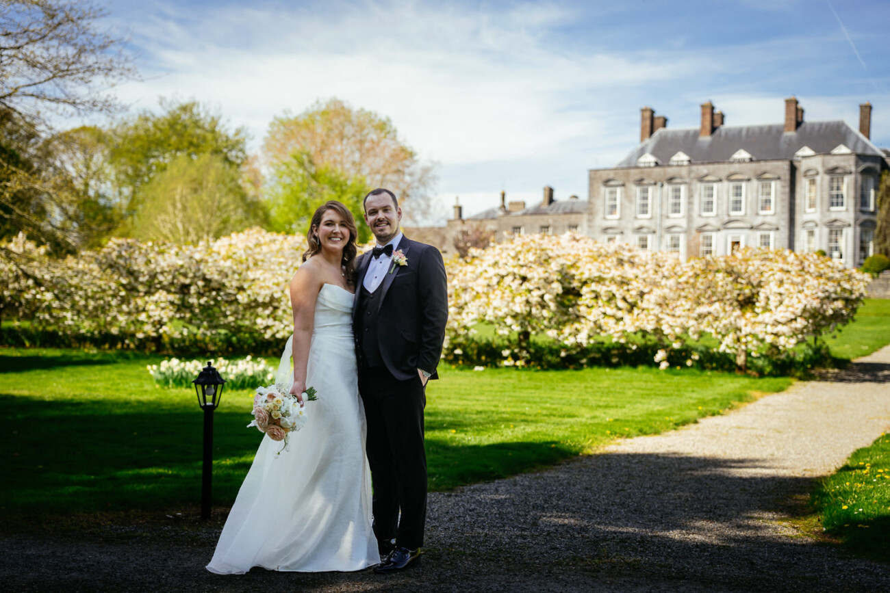 Why Your Photographer Won't Give You the RAW Files (And Why That's Perfectly Reasonable) 2 A bride and groom stand together outdoors on a gravel path, smiling, with a large historic building and flowering shrubs in the background.