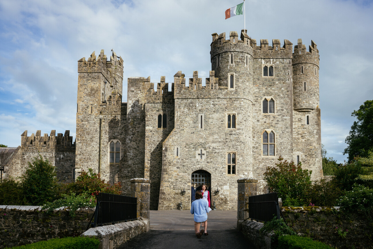 Two people walk towards Kilkea Castle with its battlements and towers, an Irish flag flying above—an enchanting setting perfect for a proposal or engagement shoot.